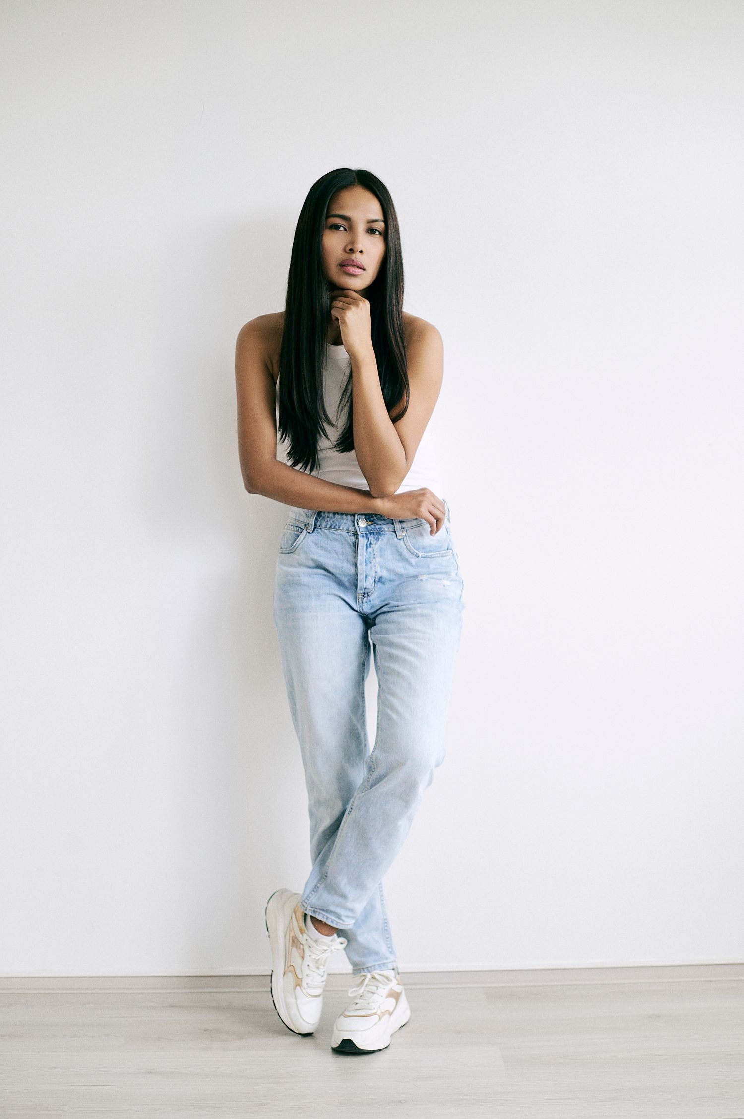 Young woman in light blue jeans and white tank top posing against a white wall wearing chunky sneakers.