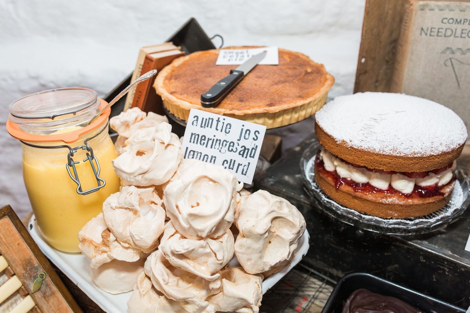 Assorted homemade baked goods including meringues, lemon curd, fruit tart, and Victoria sponge cake displayed on a market stall.
