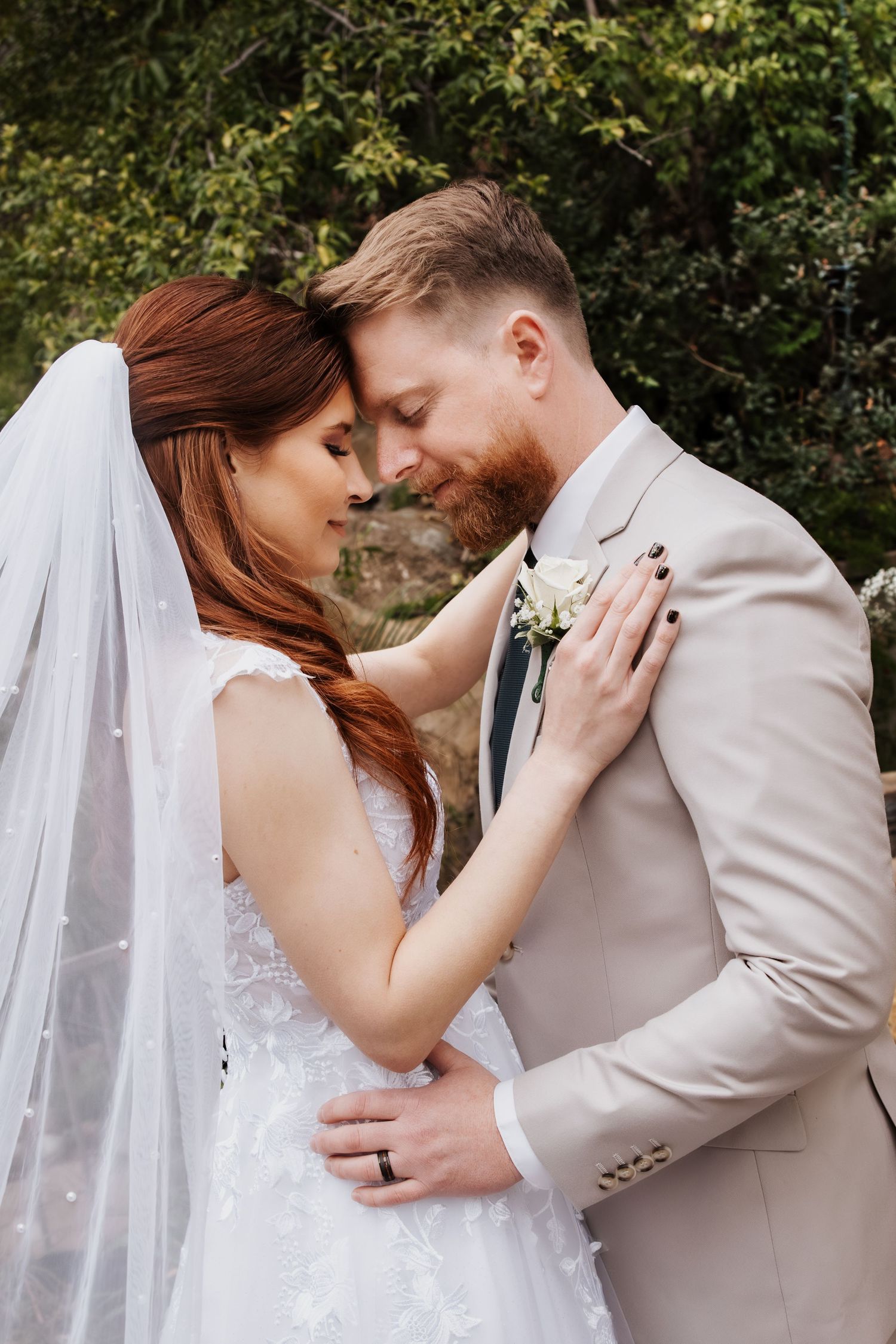 Bride with red hair in lace gown and groom in beige suit share tender forehead touch outdoors.