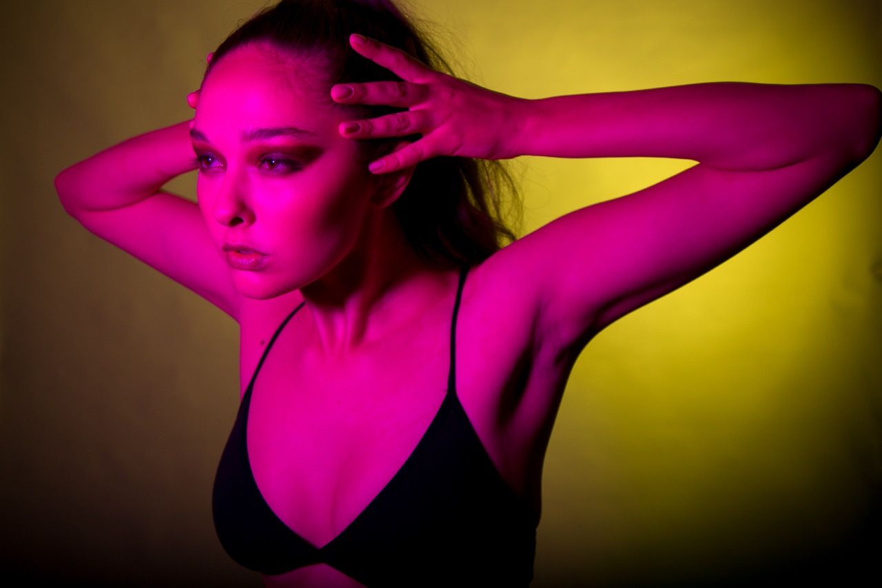 Woman posing in black bralette under vibrant pink and yellow neon lighting in a dramatic studio setting.