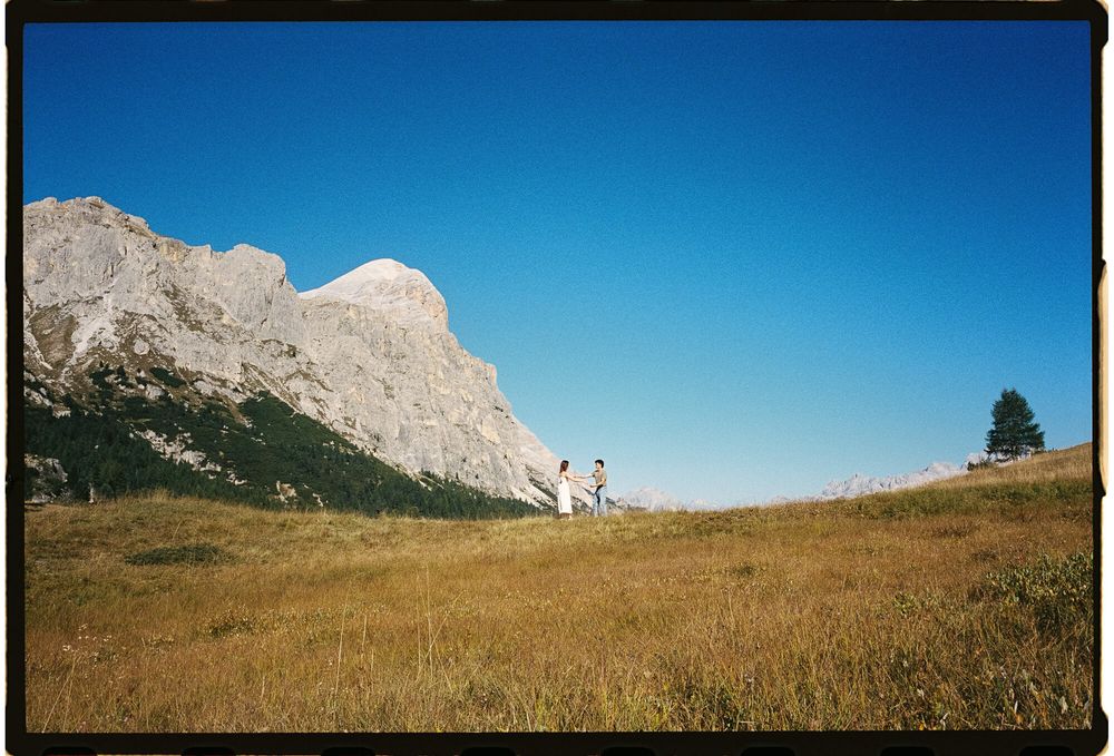 Two hikers walk across a golden meadow beneath dramatic rocky mountain peaks under a clear blue sky.