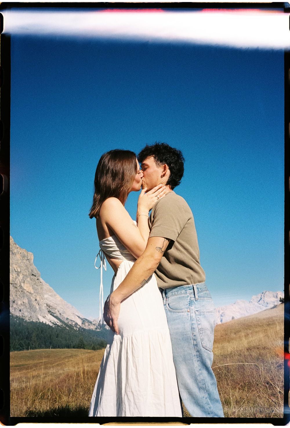 A couple sharing a romantic kiss in a scenic mountain meadow under a clear blue sky, woman in white dress.