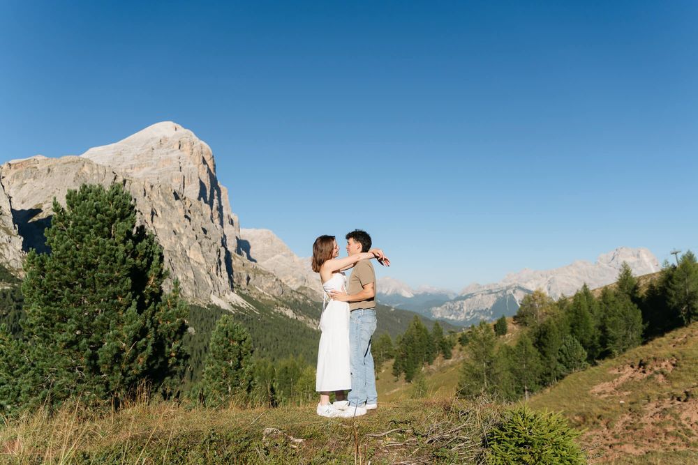 Romantic couple kissing on a scenic mountain hillside with dramatic rocky peaks and blue sky.