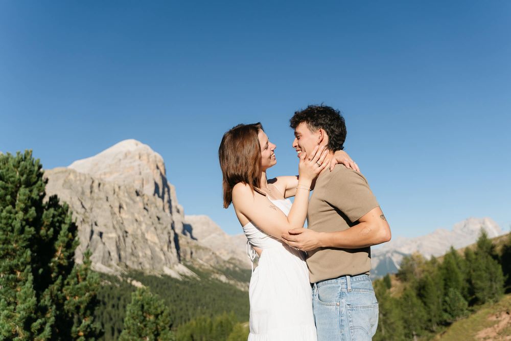Couple embracing in the Dolomites with rocky mountains and green trees under a clear blue sky.
