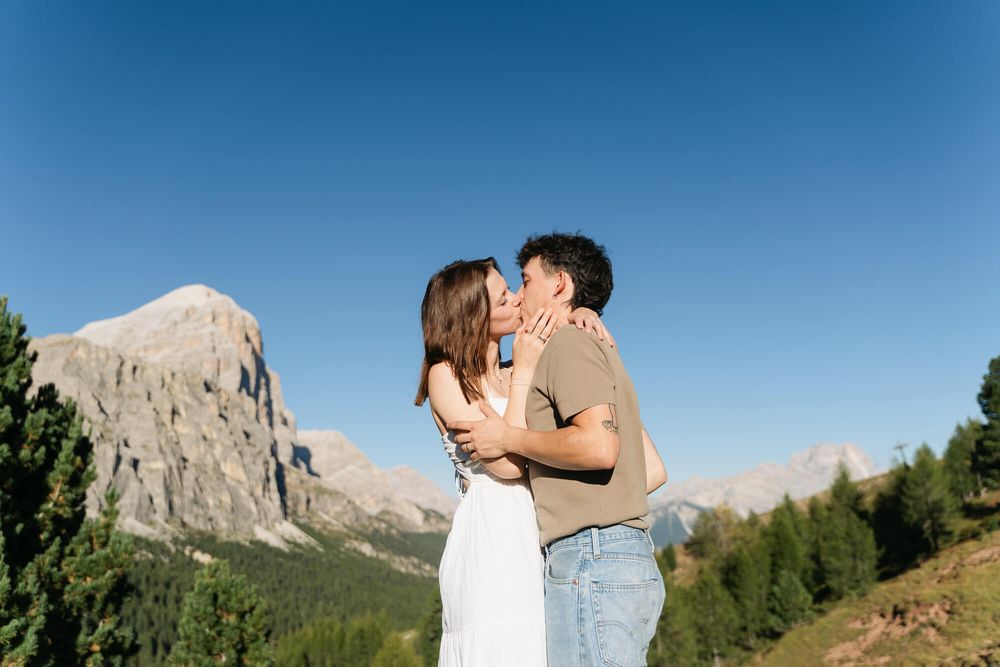 Couple kissing in the Dolomites mountains with pine trees and rocky peaks under a clear blue sky.