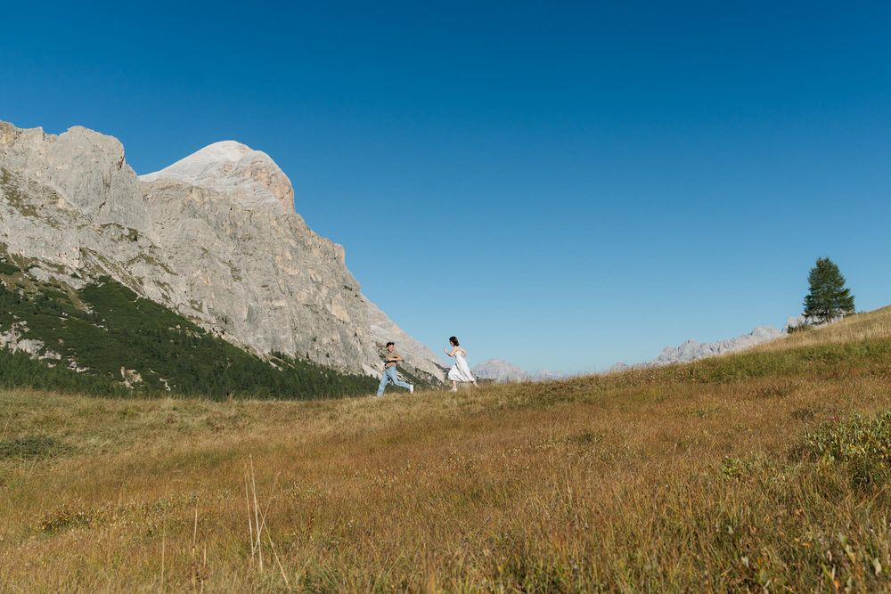 Couple walking hand in hand on a grassy alpine meadow with dramatic rocky mountain cliffs under a clear blue sky.