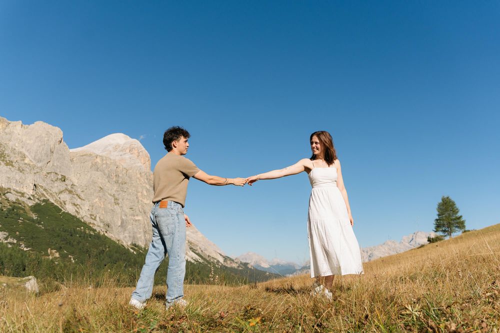 Couple holding hands in a green alpine meadow with rocky mountains and clear blue sky behind them.