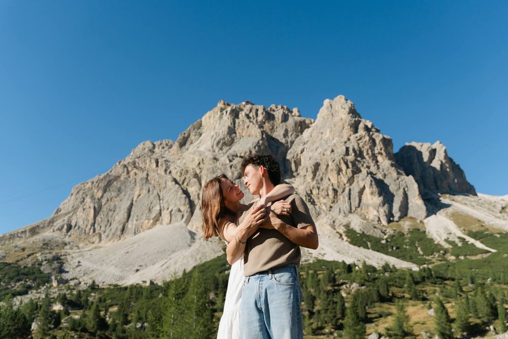 Couple embracing romantically in front of dramatic rocky Dolomites mountain peaks under clear blue sky.