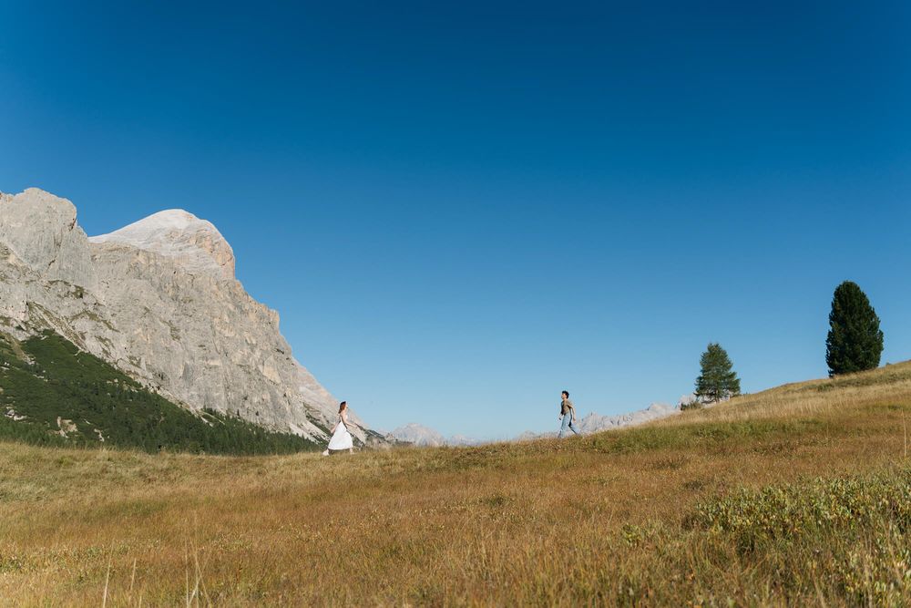 Bride and groom walk across a golden meadow with dramatic rocky mountain peaks and clear blue sky in the Dolomites.