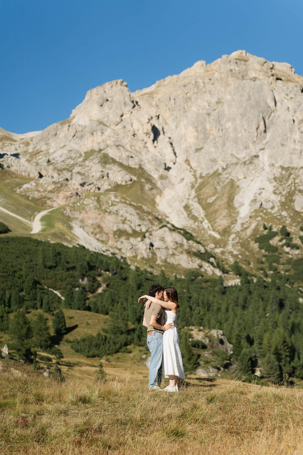 Couple embracing on a mountain meadow with dramatic rocky peaks and pine forests under a clear blue sky.