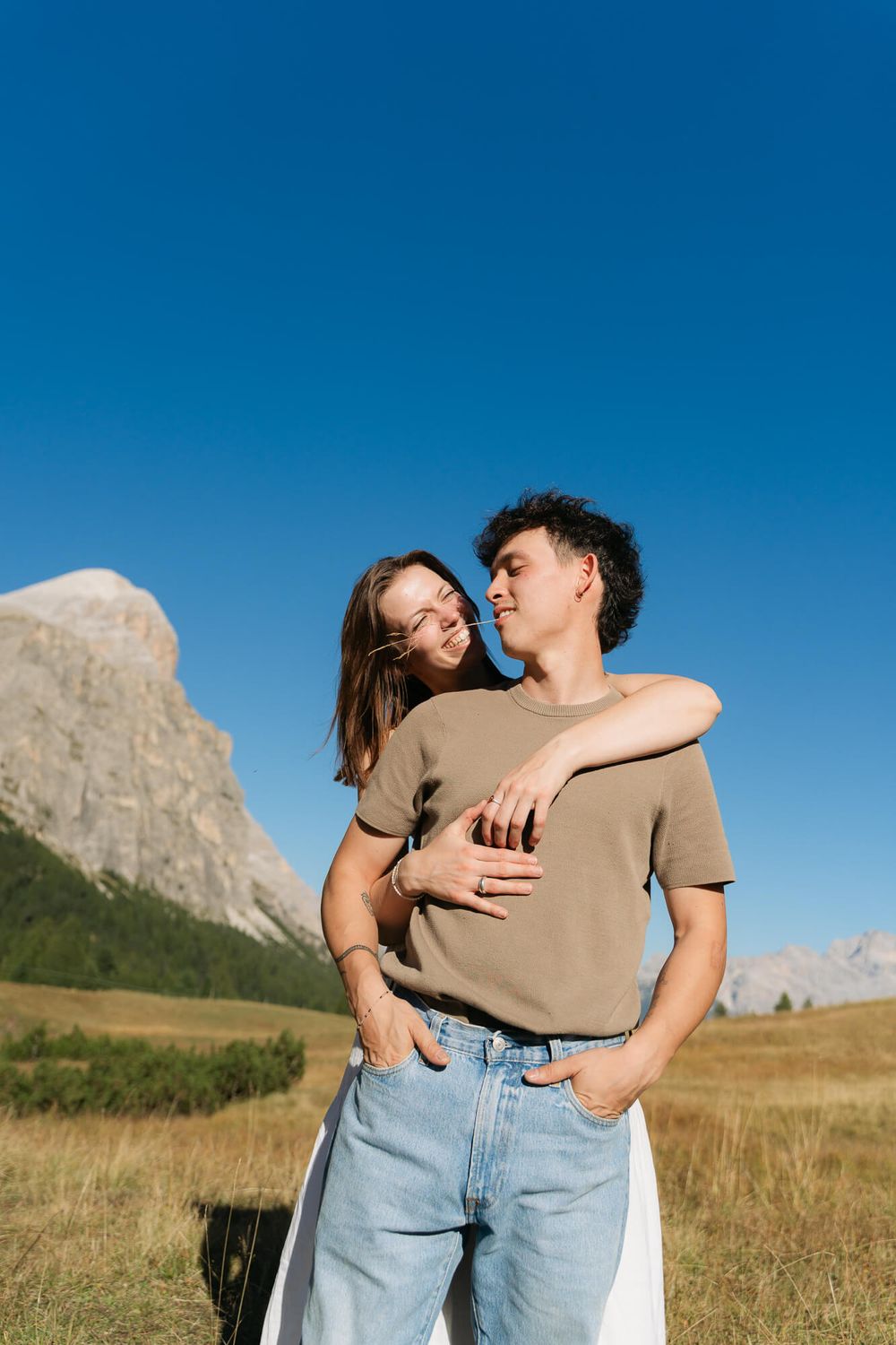 Couple embracing in a mountain meadow under a clear blue sky, with rocky peaks and green trees in the background.