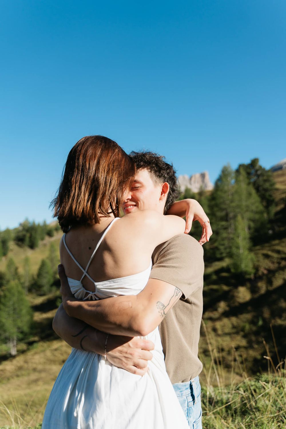 Couple embracing in a mountain meadow with green trees and blue sky, woman in white dress hugging smiling man outdoors.