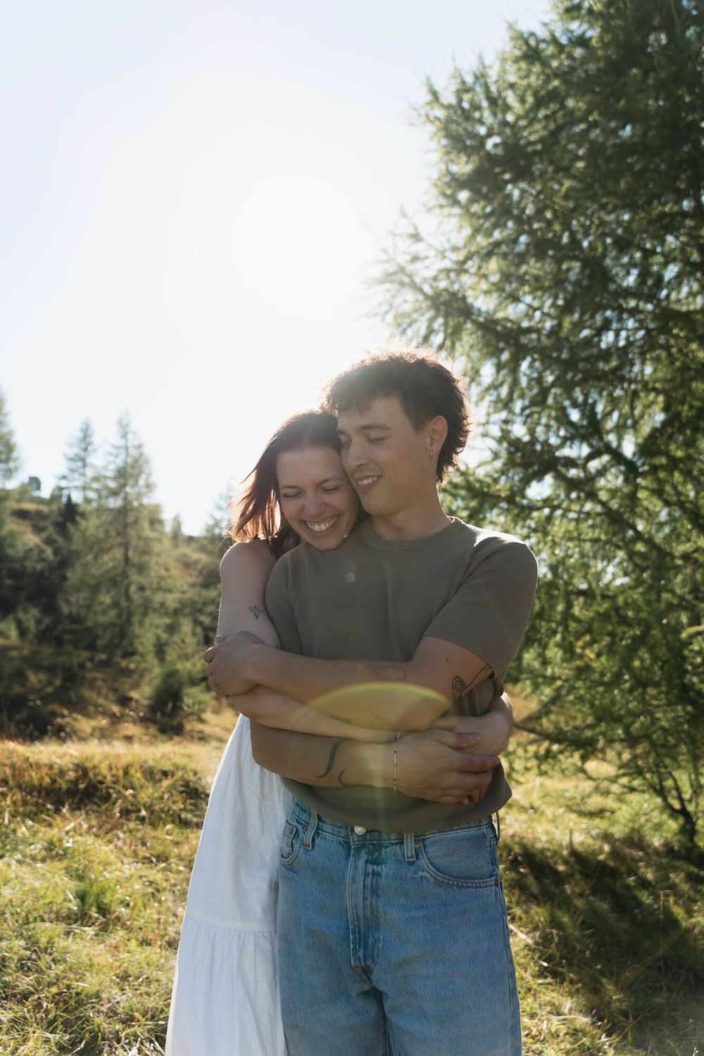 Happy couple embracing outdoors in sunny meadow with trees, wearing casual clothes, sharing a joyful moment in nature.