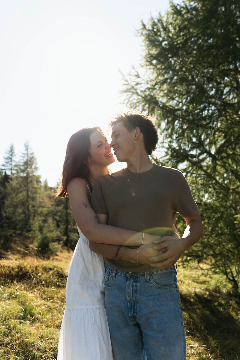 A couple embraces and nearly kisses outdoors in a sunny alpine meadow with green trees in the background.