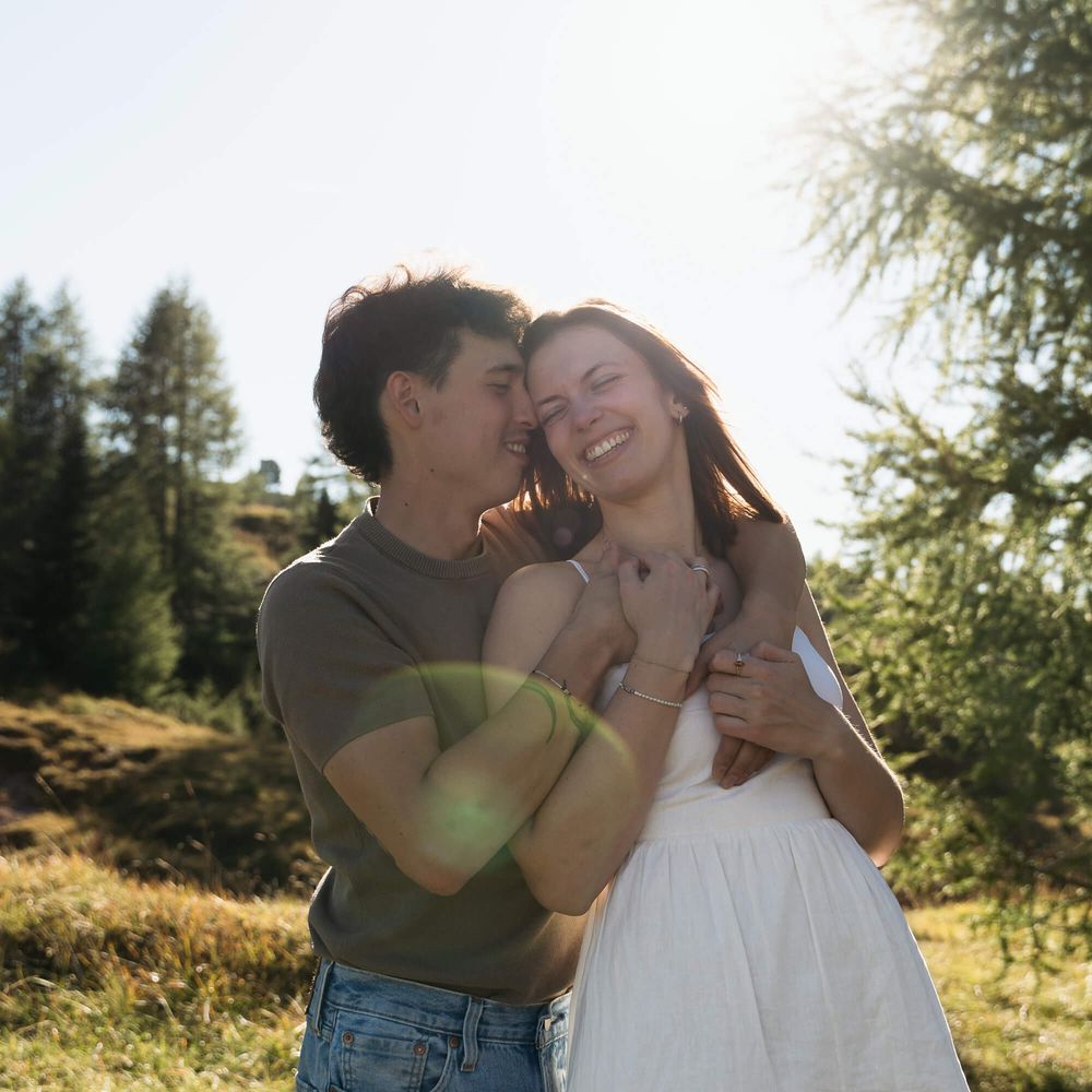 Romantic couple hugging in sunny meadow with man kissing woman's cheek near pine trees.