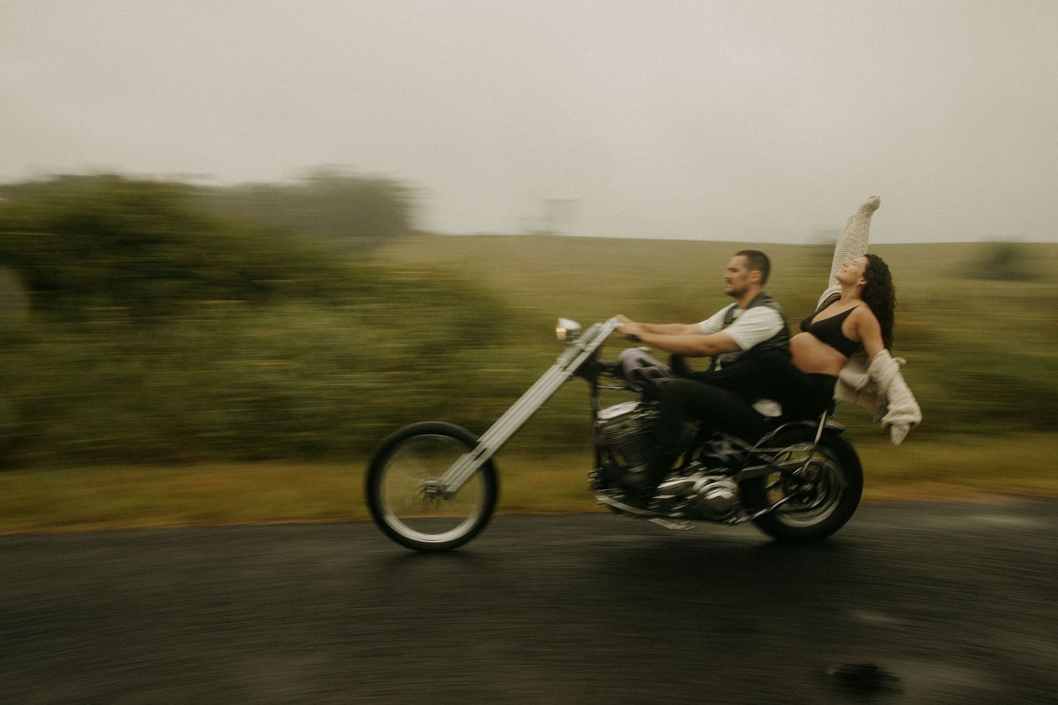 A couple rides a chopper motorcycle on a rainy road, the bride raising her arm joyfully in a blur of motion.