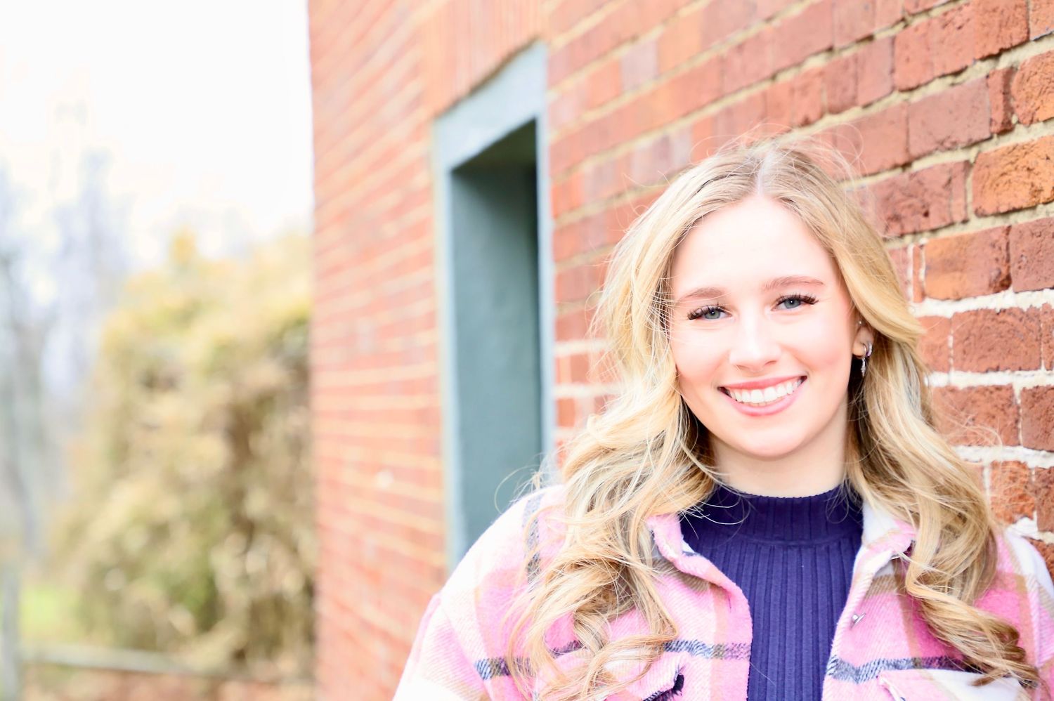 Smiling blonde senior wearing a pink jacket beside a brick wall
