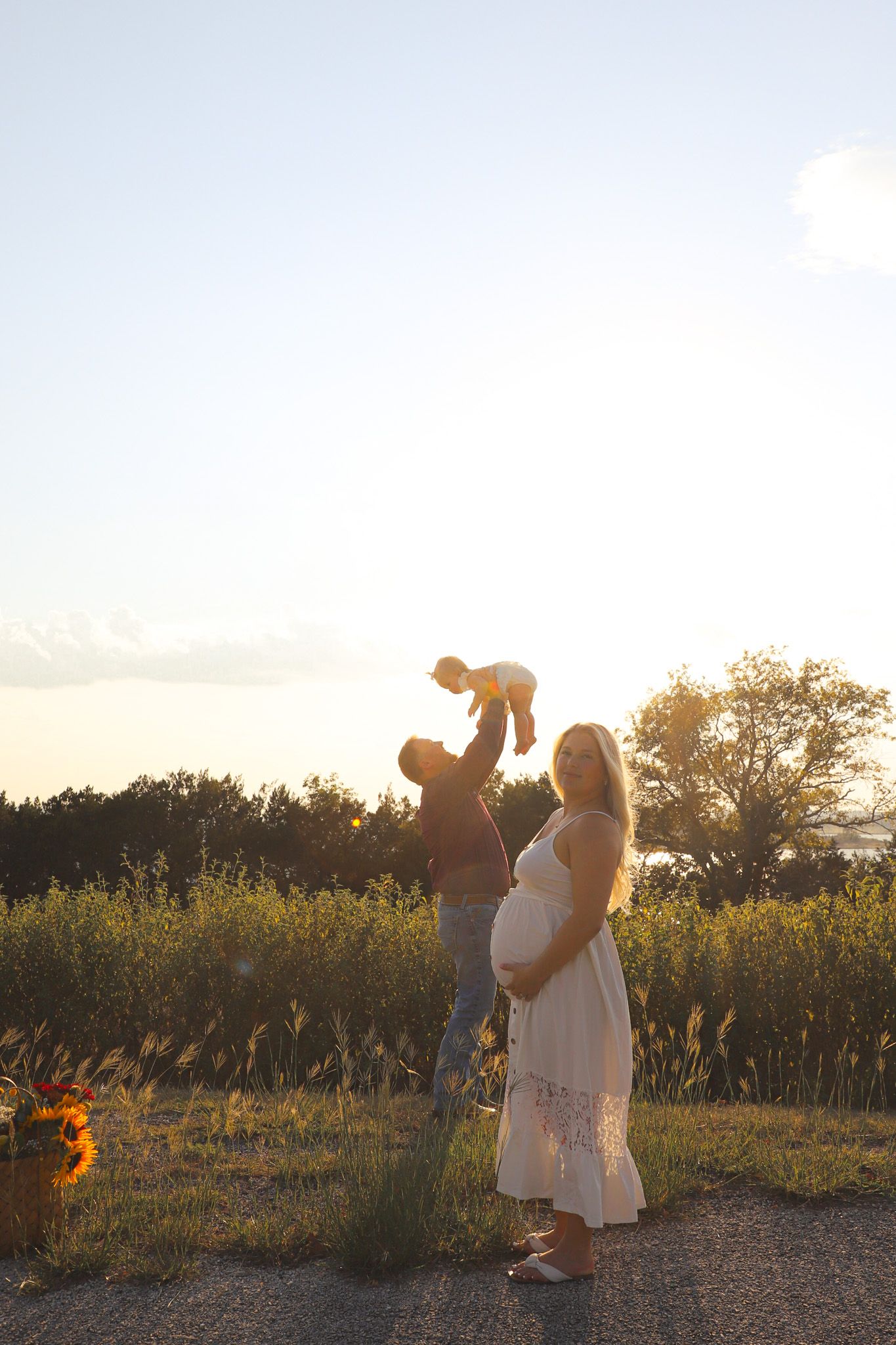 Expectant mother posing as the father lifts their baby at sunset
