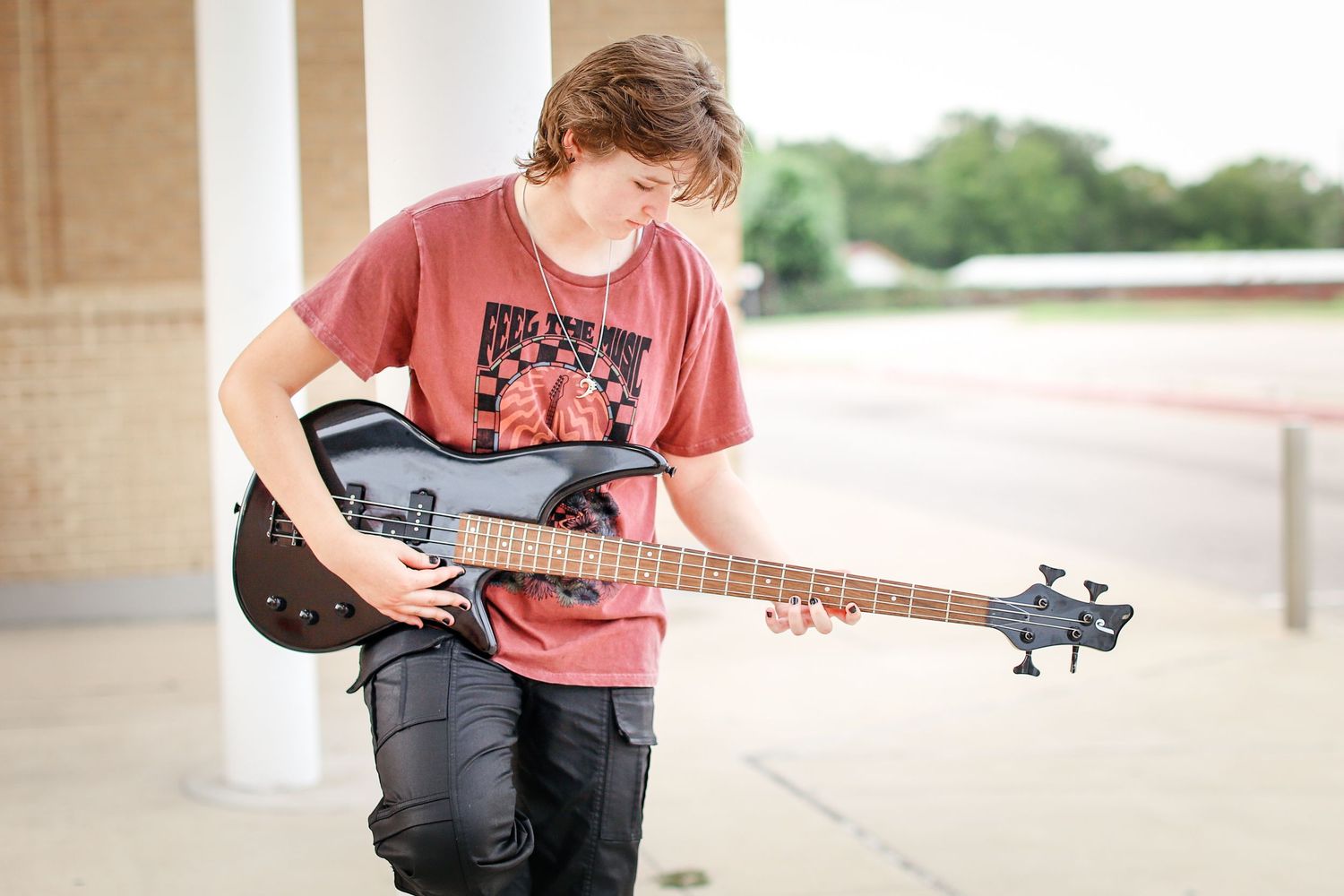 Teenage musician playing a black bass guitar outside a school