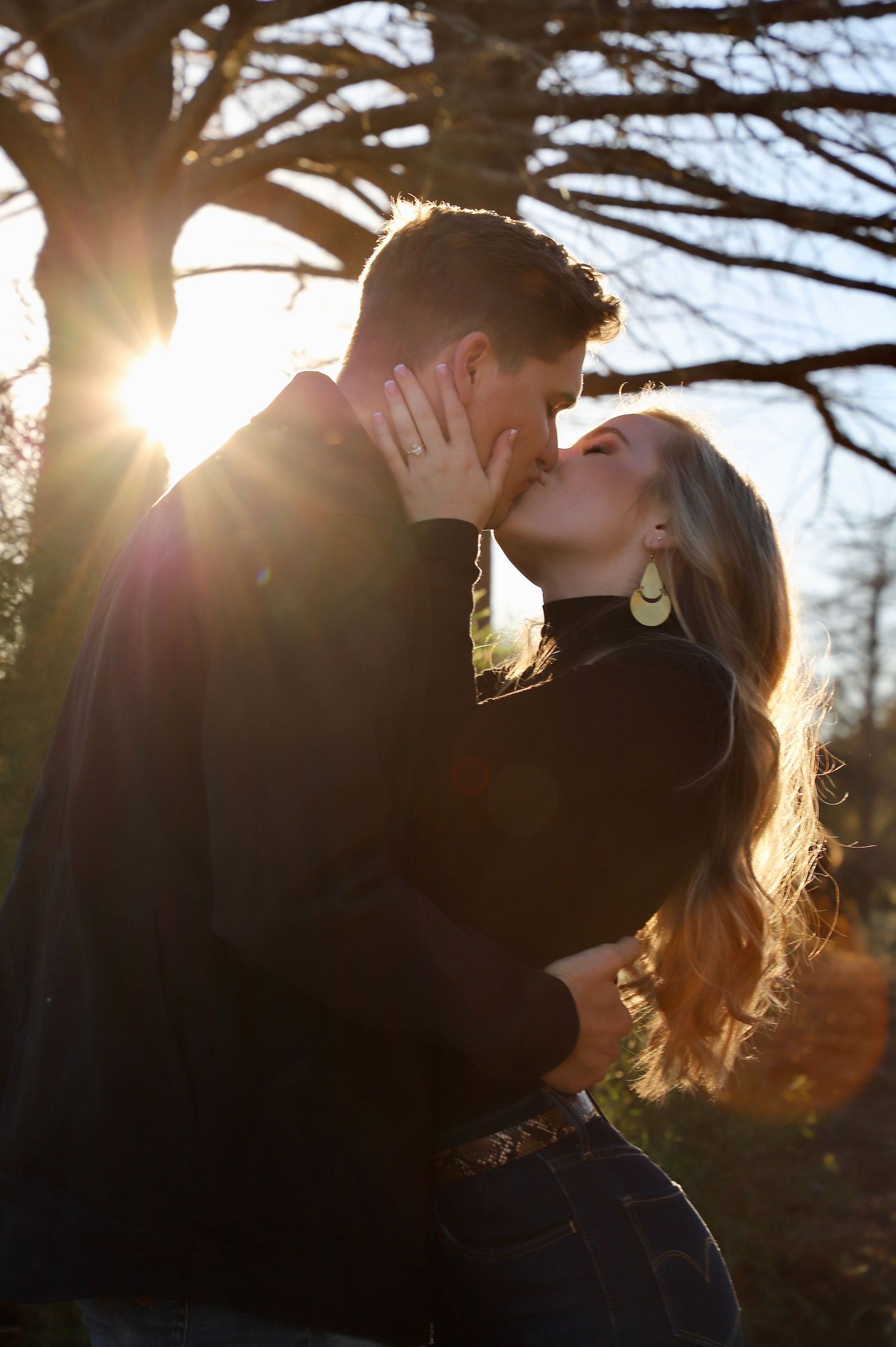 Engaged couple kissing beneath a large tree at sunset
