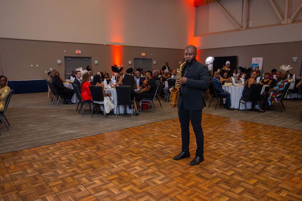 Event speaker addresses attendees in formal attire at banquet hall with orange lighting and parquet dance floor.