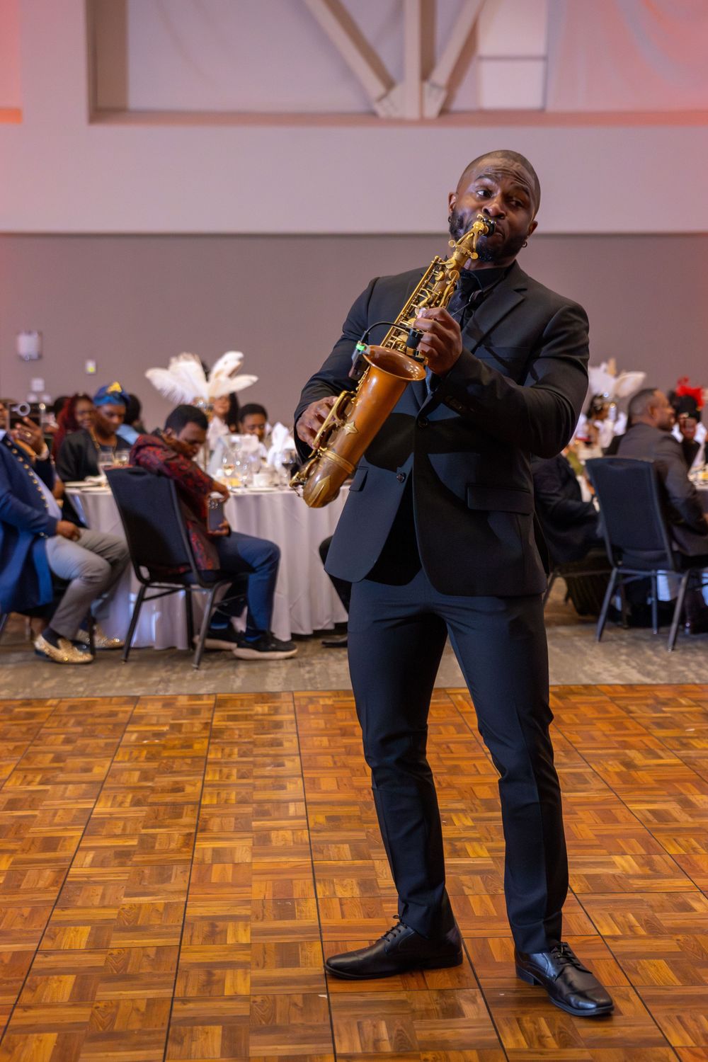 A saxophonist in a dark suit performs energetically on a parquet dance floor at an indoor event.
