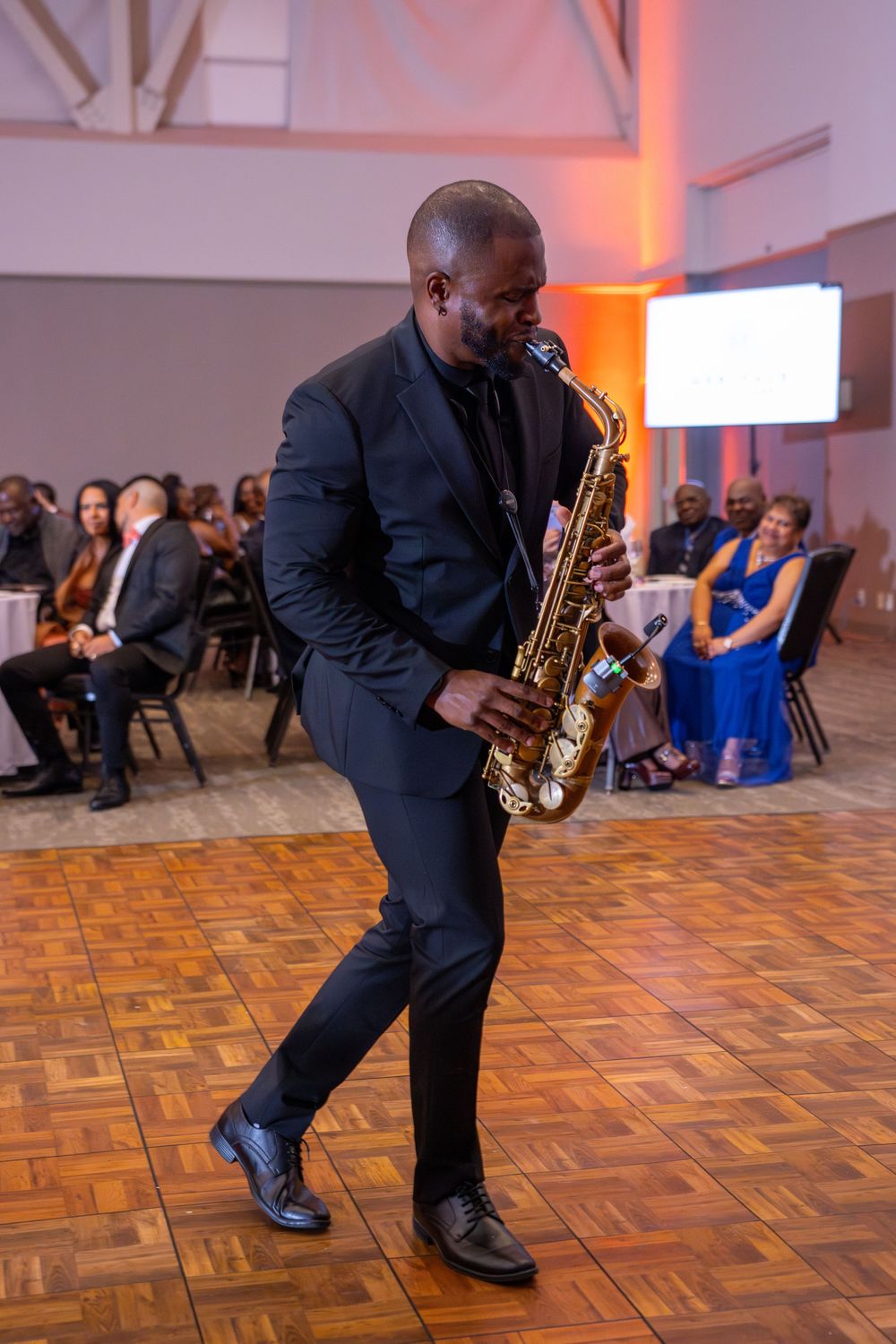 Saxophone performer in dark suit plays energetically on a parquet dance floor with orange ambient lighting.