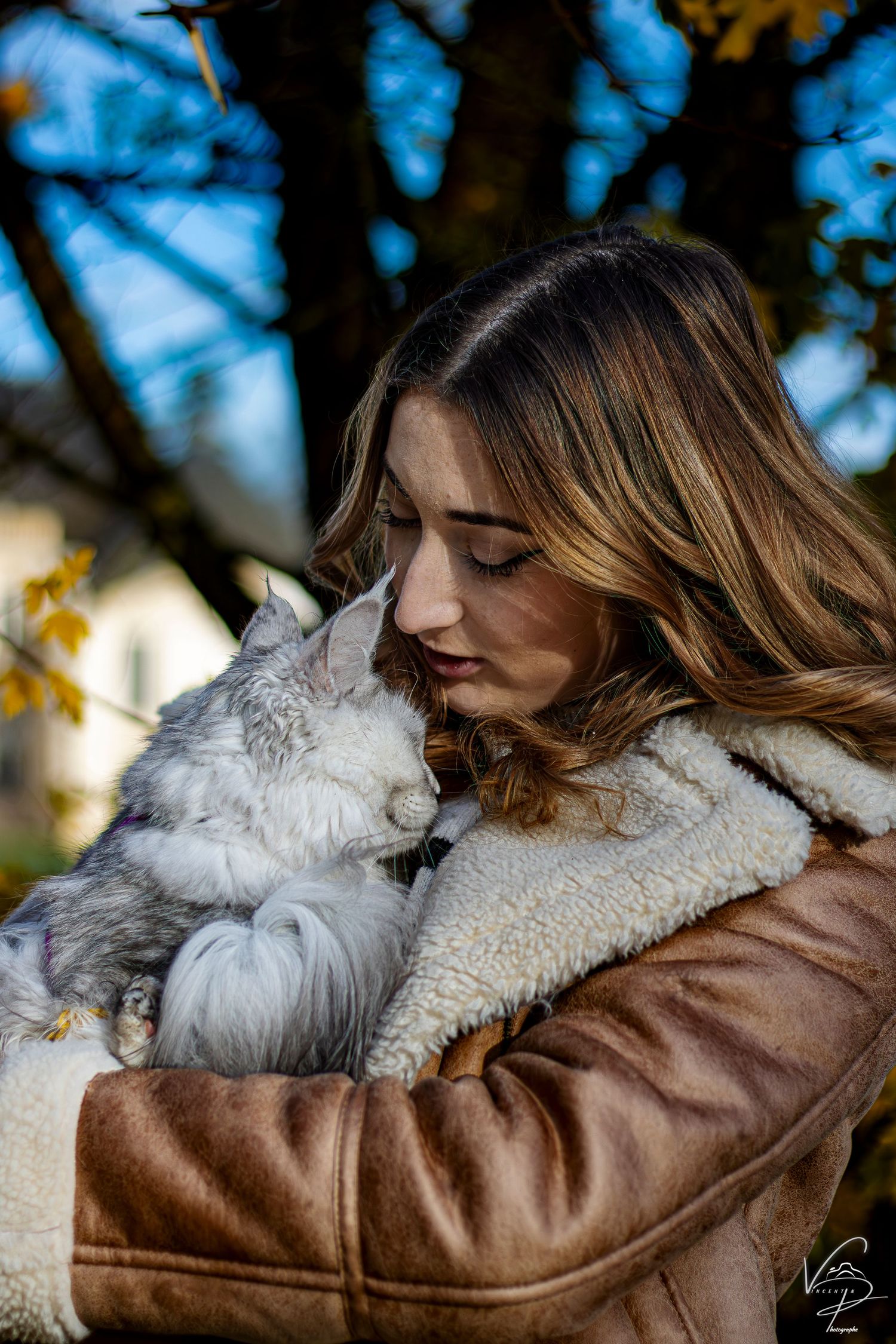 A fluffy white cat snuggles against someone wearing a brown leather jacket in an outdoor setting.