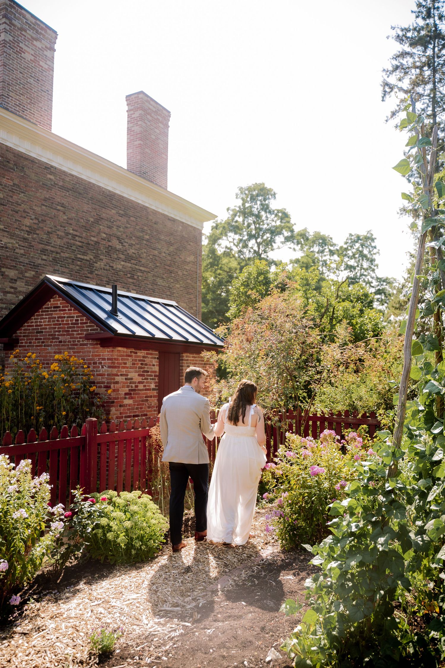 A couple walks along a garden path beside a brick building with flowering bushes and a red fence on a sunny day.