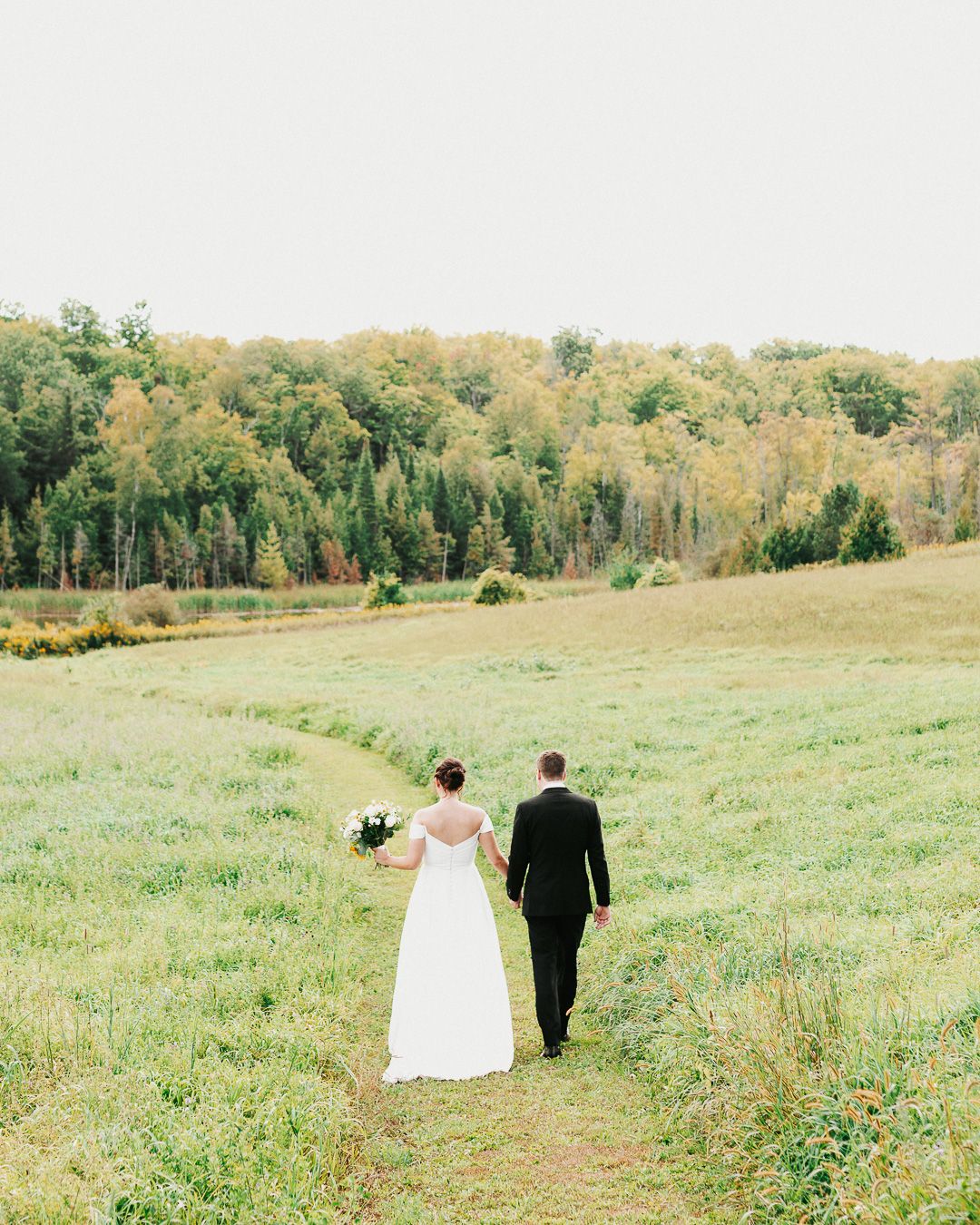 A couple walks down a grassy path through rolling hills with autumn trees in the background.