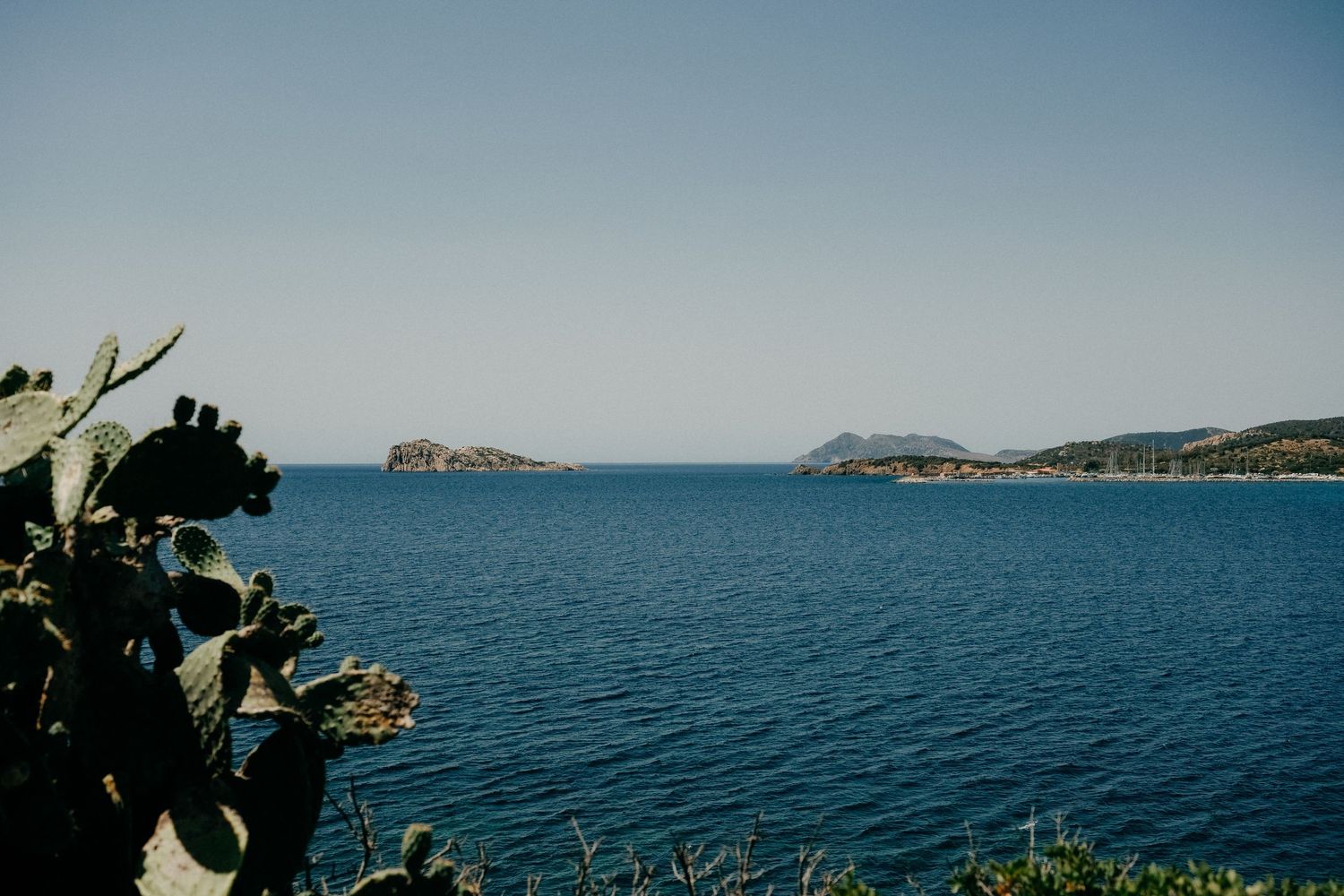 Scenic Mediterranean coastline with deep blue waters and distant mountains visible through lush green foliage.