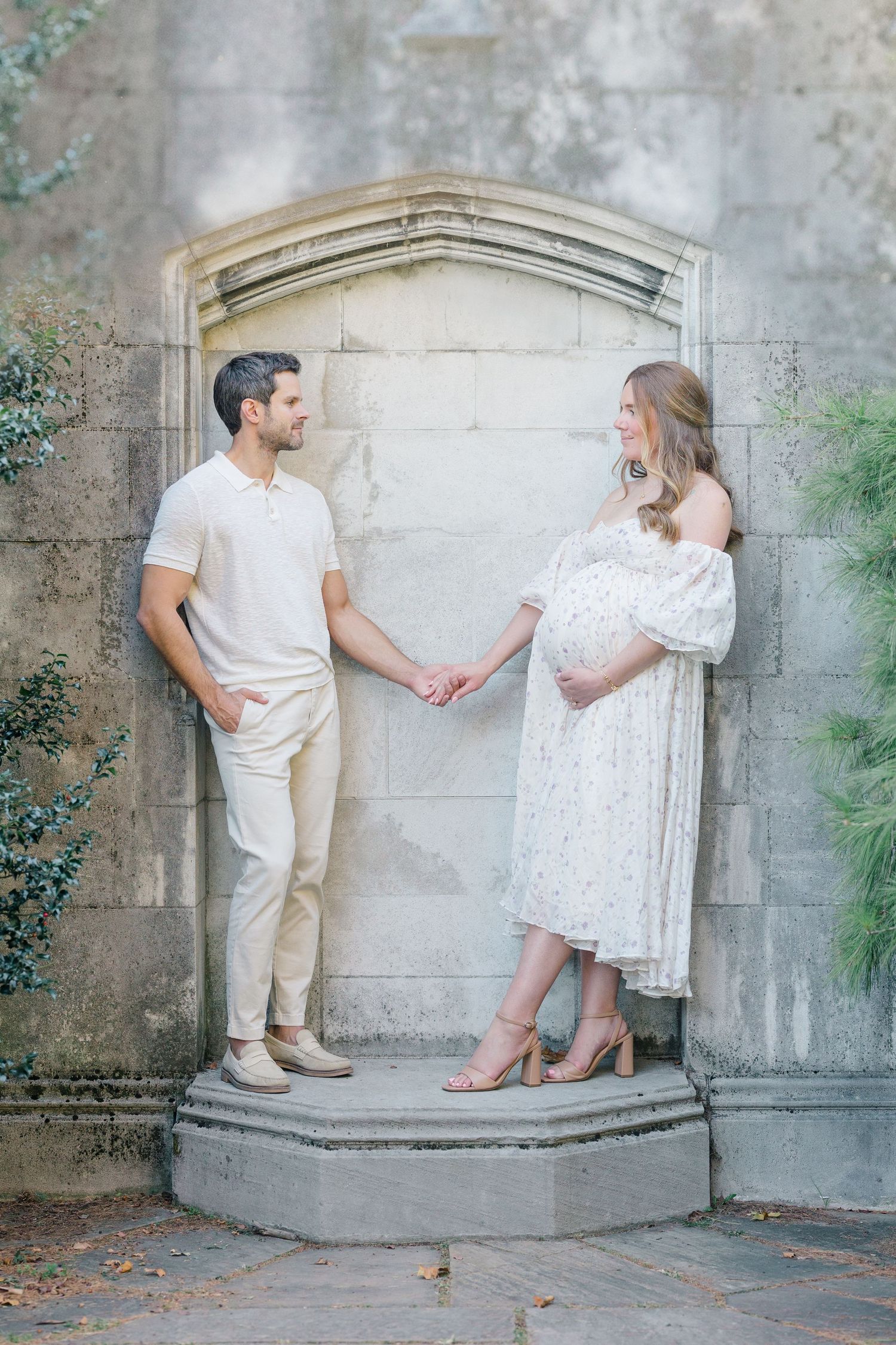 A couple in white outfits playfully interacts against a stone wall in a romantic outdoor photo series.
