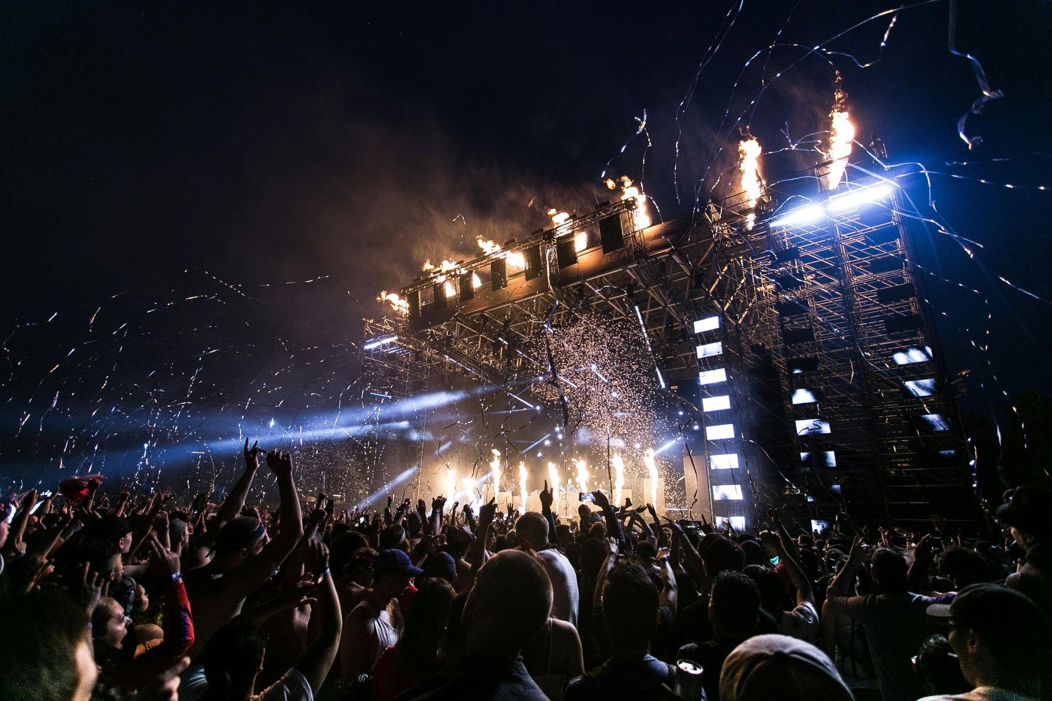 High-energy night music festival with cheering crowd, bright stage lights, fire effects, and confetti creating an electric live concert atmo