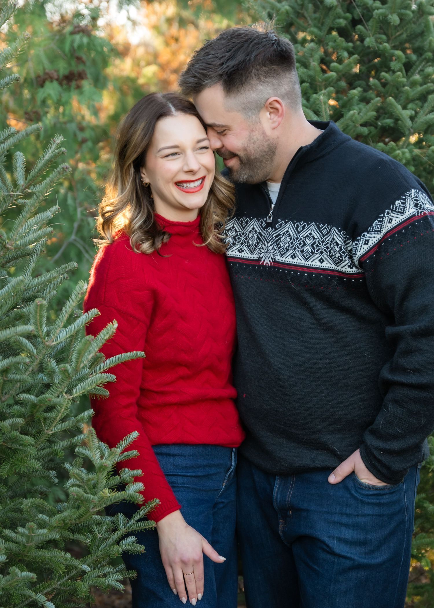A couple in festive sweaters share a joyful moment among evergreen trees during a holiday photo session.