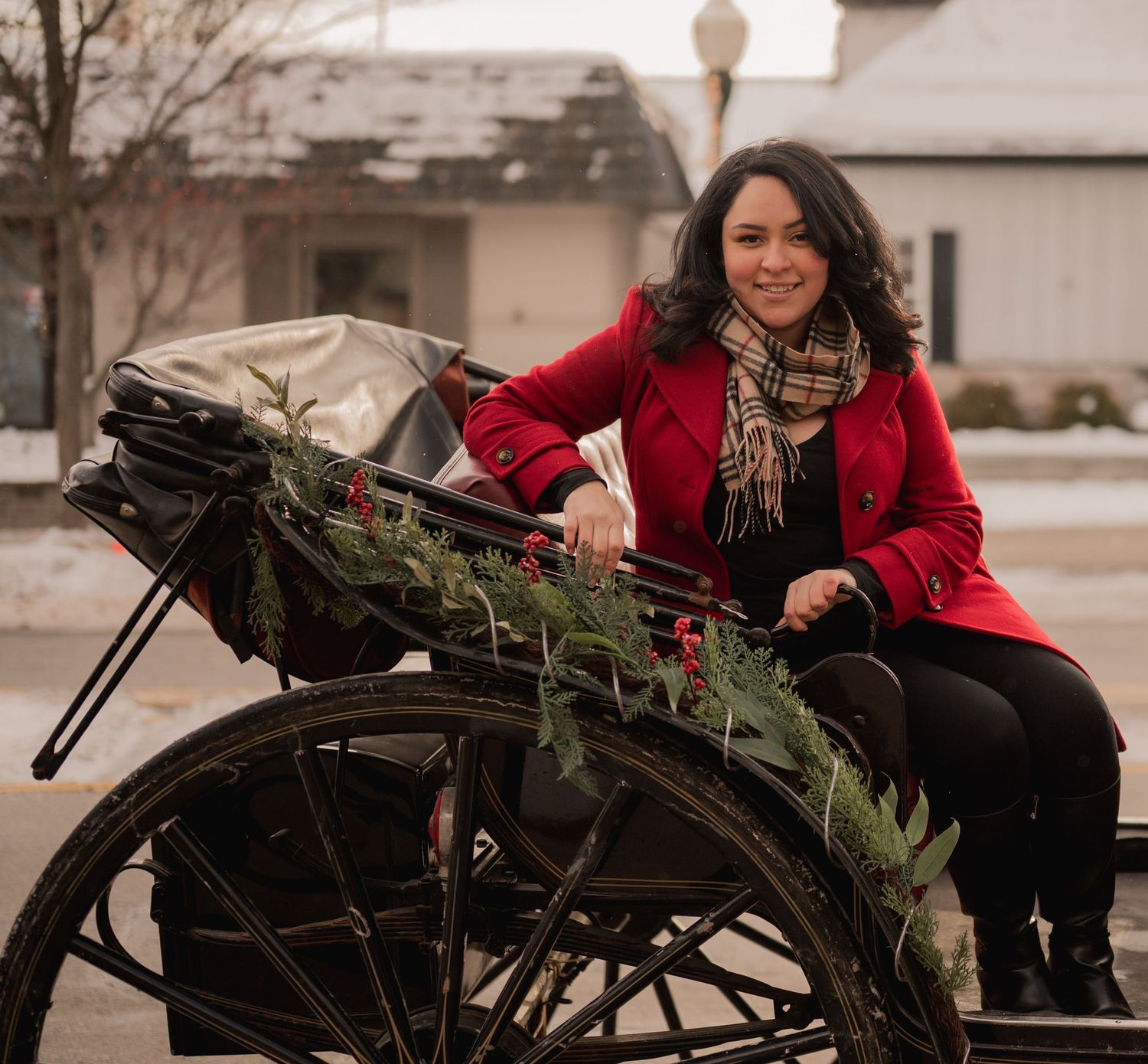 A person in a red coat sits in a vintage carriage decorated with holiday greenery on a winter day.