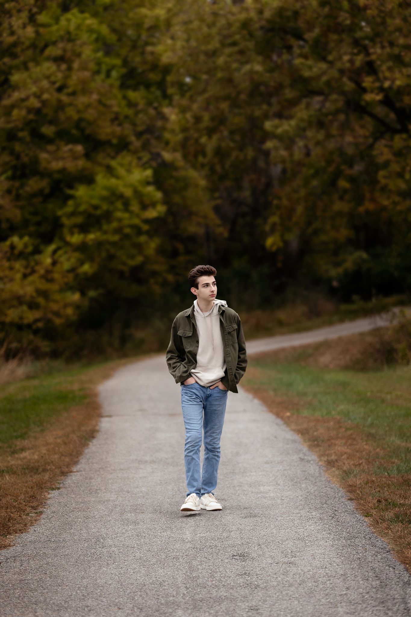 A person in a dark jacket and jeans walks alone on a gravel path surrounded by autumn trees.