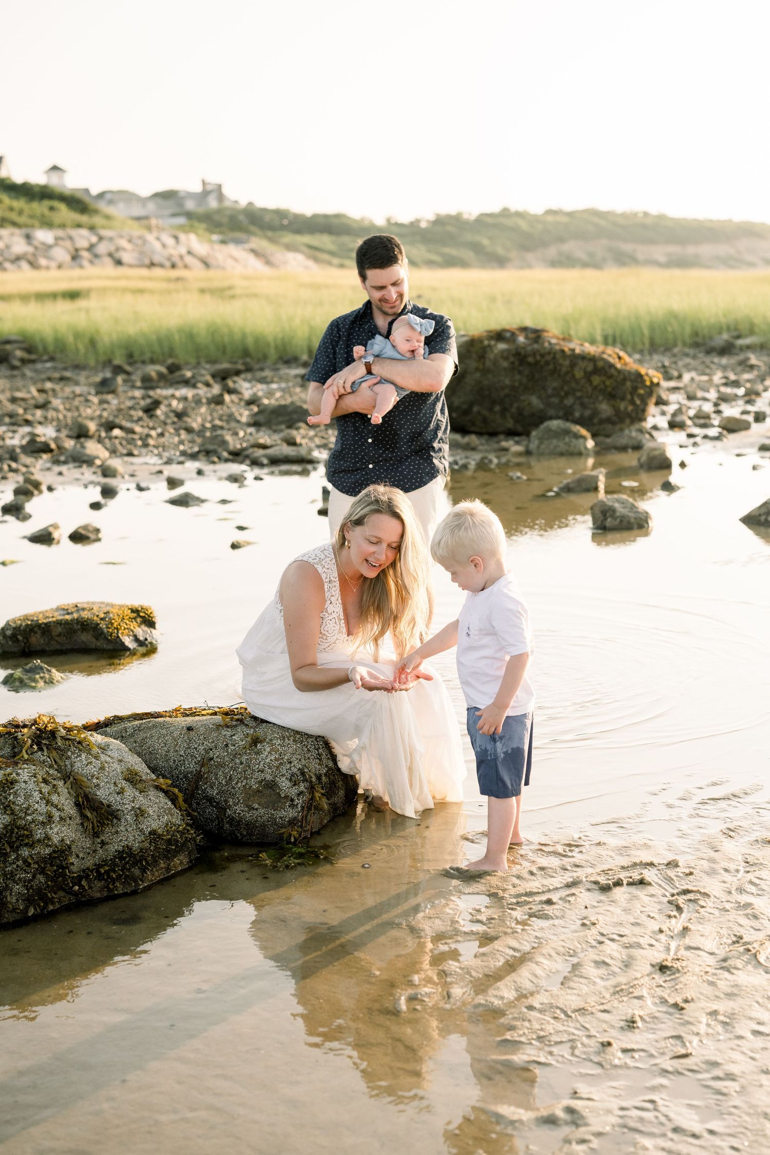 Family in white outfits exploring tide pools and rocks along a scenic beach at sunset.