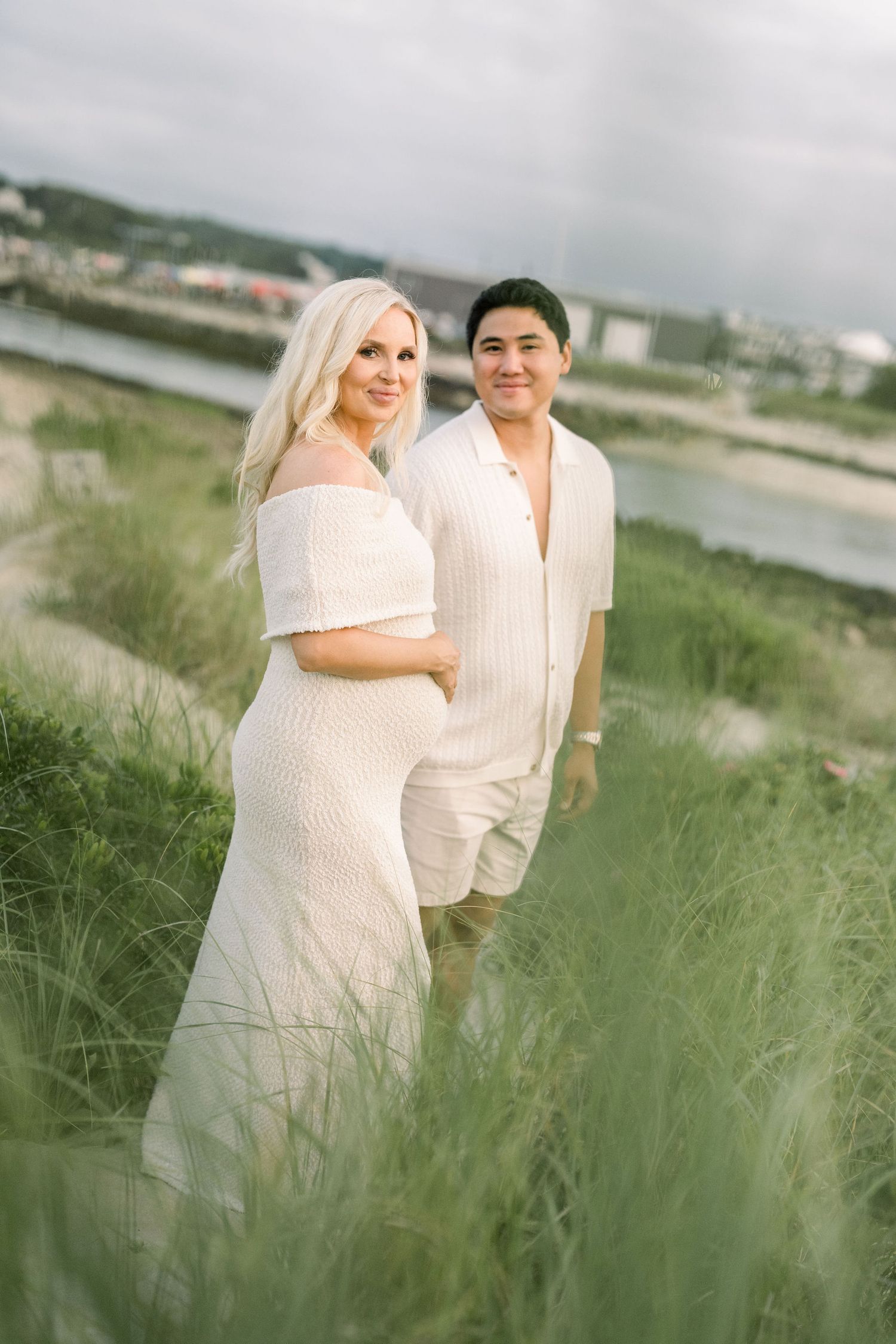 Couple in white outfits standing together in tall grass by a waterfront on a cloudy day.