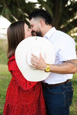 Couple kissing behind cowboy hat under tree during golden hour.