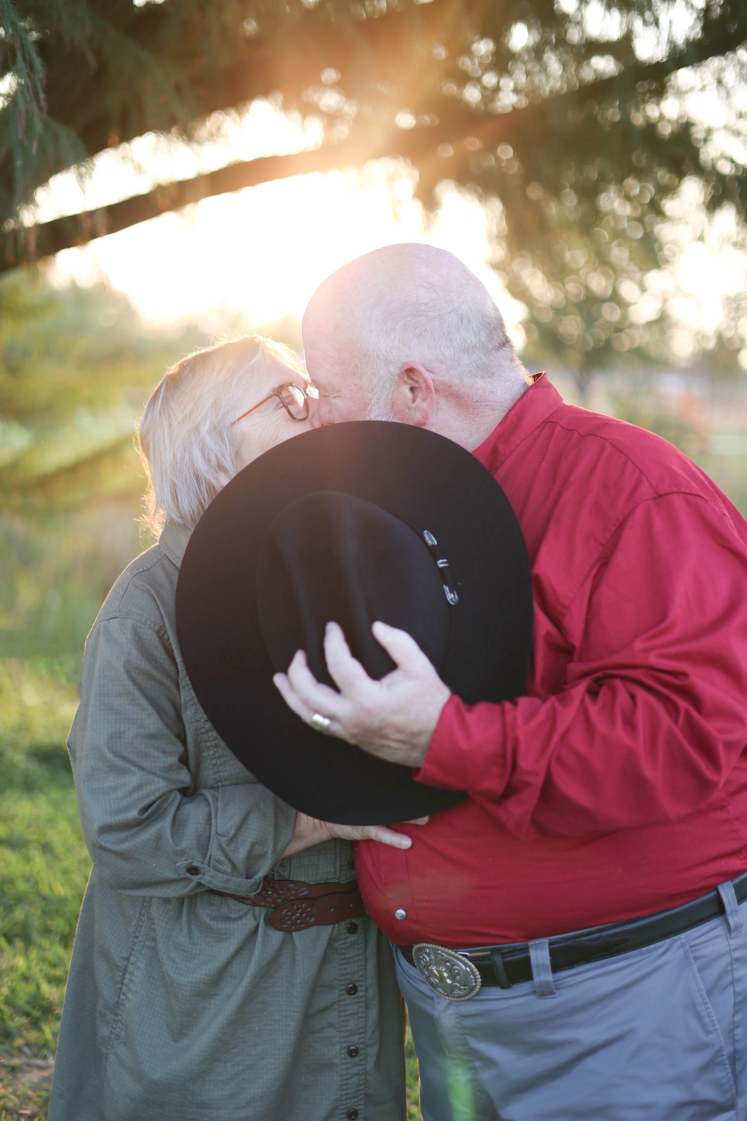 Couple kissing behind cowboy hat under tree during golden hour.