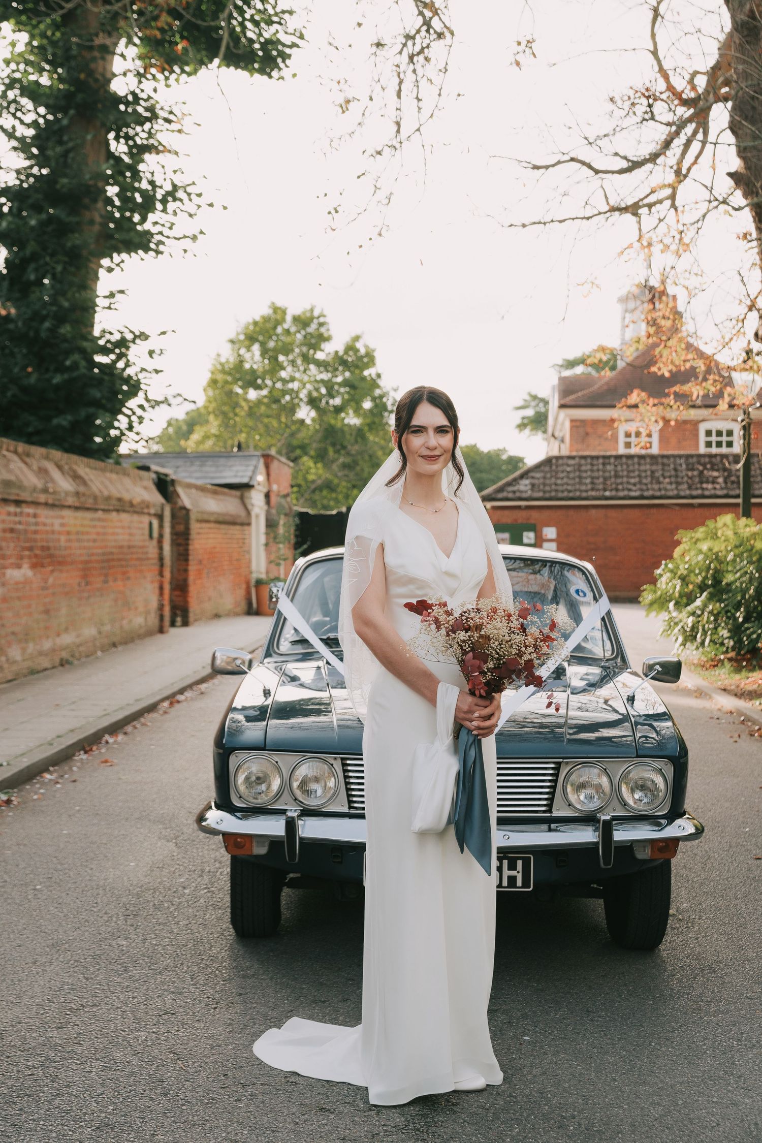A vintage Rolls Royce parked on a brick-lined street with someone in a white wedding dress standing beside it.