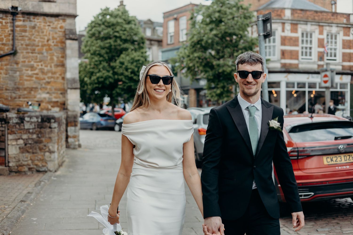 A couple in formal wedding attire walks together and smiles during an outdoor photo session in an urban setting.