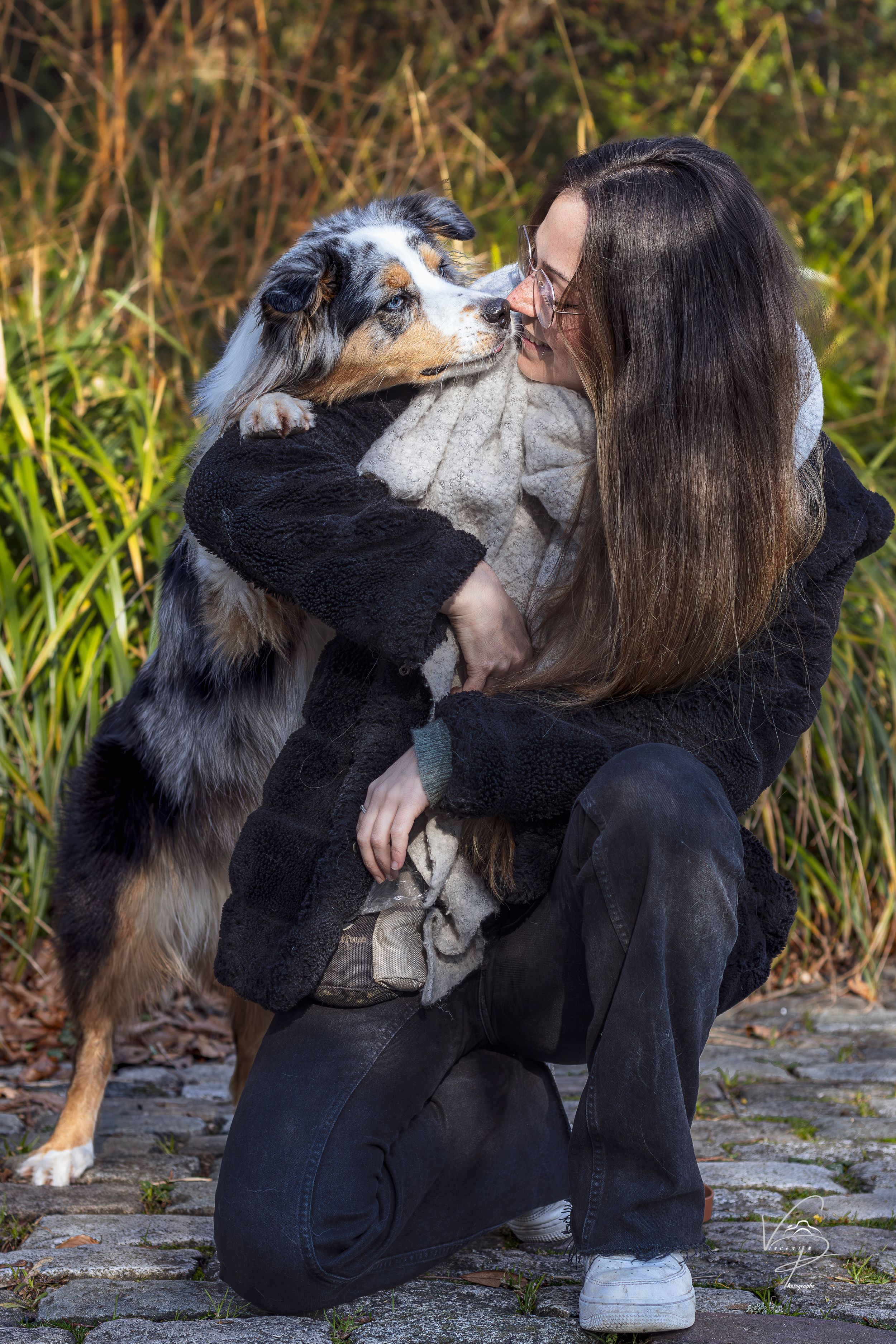 A young woman crouches on a cobblestone path, hugging an Australian Shepherd dog outdoors.