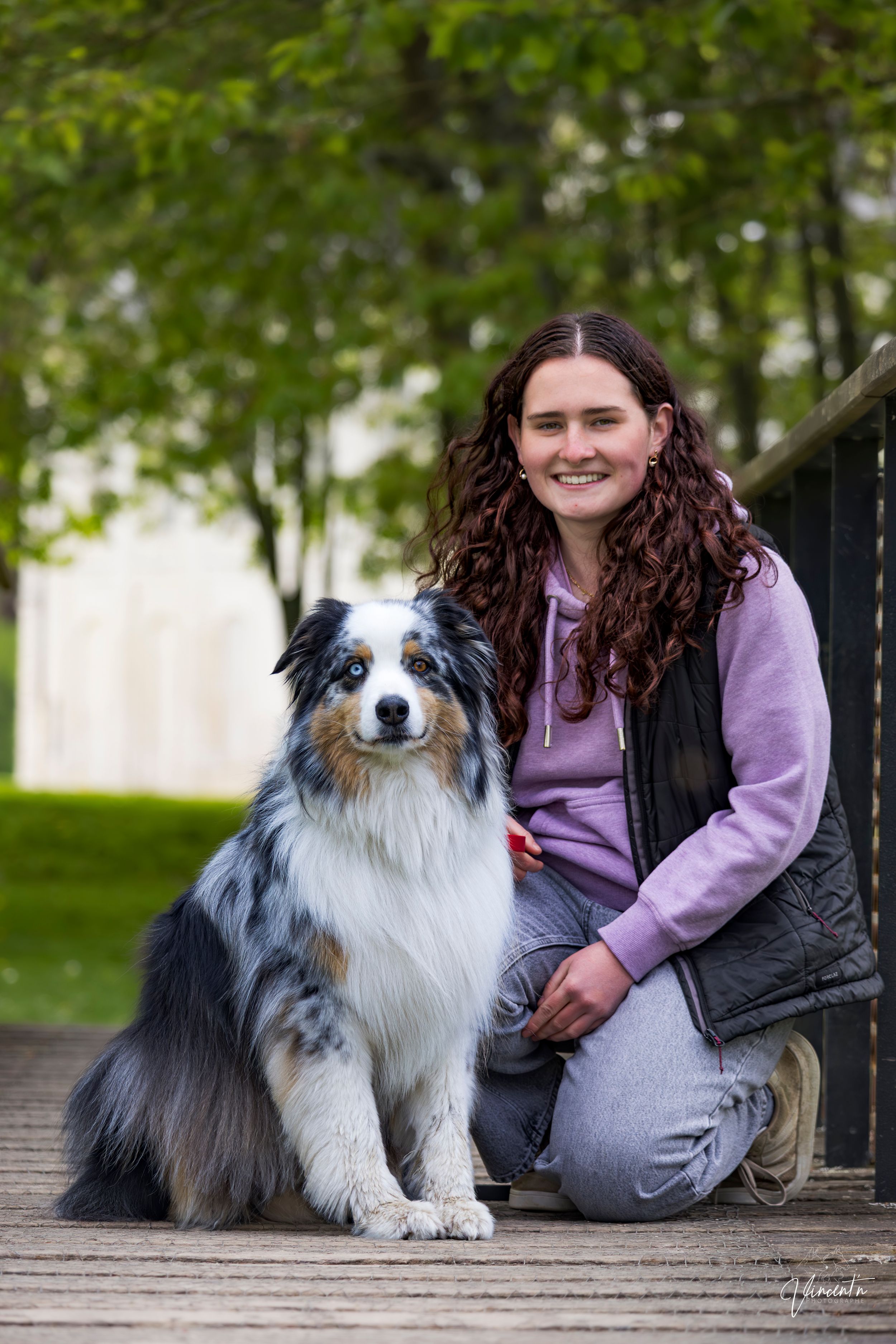 Smiling young woman with curly hair posing with her blue merle Australian Shepherd on a wooden deck.