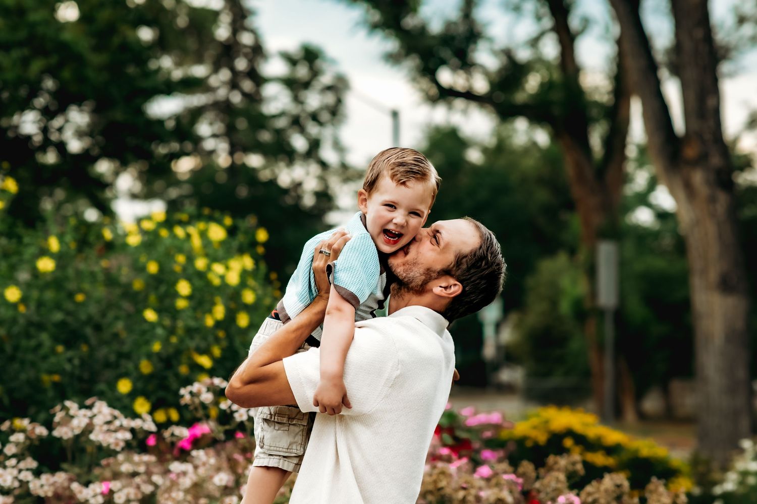 A couple shares a romantic embrace in a garden setting surrounded by blooming flowers and trees.