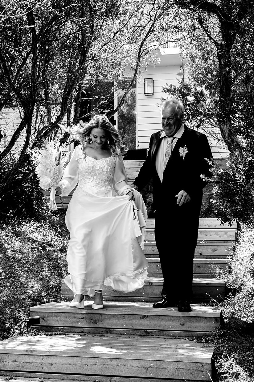 A bride and her escort descend wooden steps under an archway of trees in an intimate black and white photo.