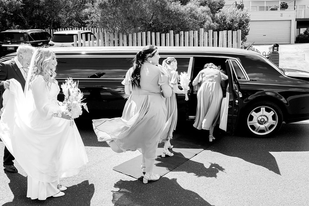 A wedding party walking near a black limousine on a sunny day, with flowing dresses catching the light.