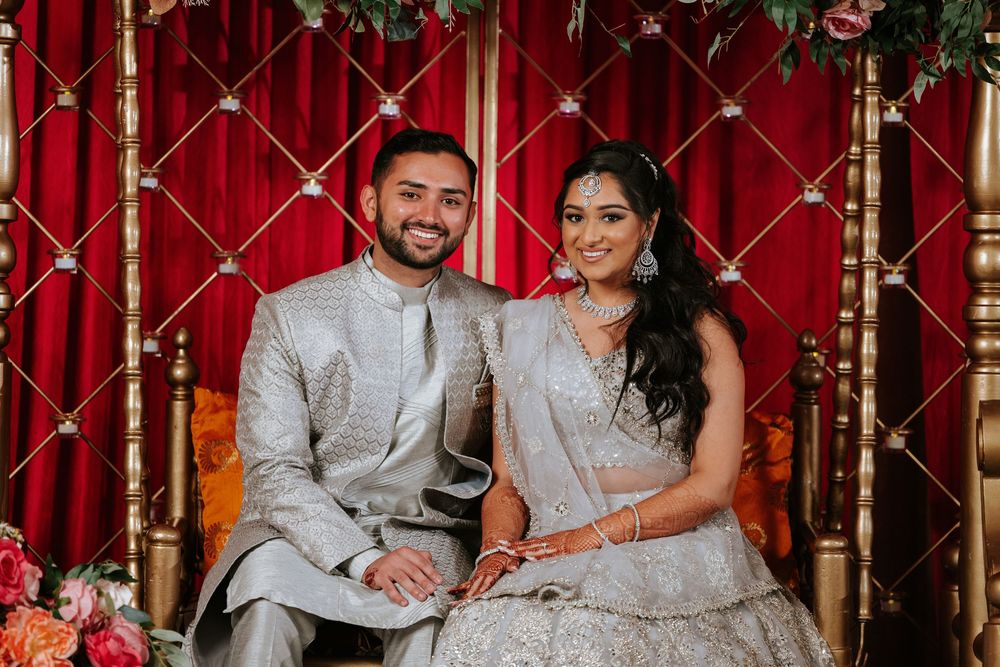A couple in elegant silver Indian wedding attire poses together against a red backdrop with decorative greenery.