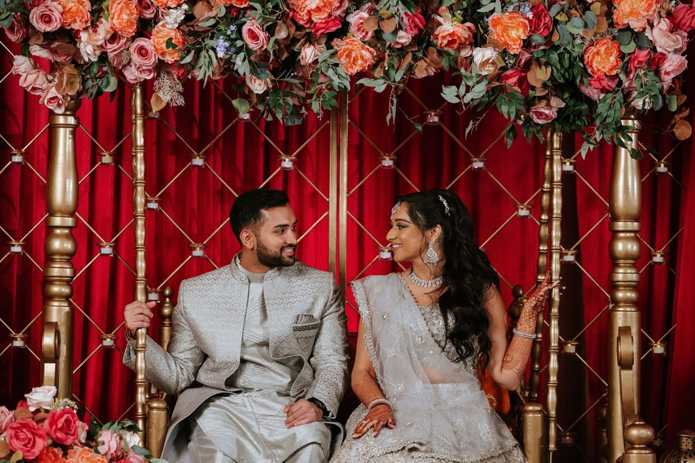 Wedding couple sits together on elegant red and gold couch surrounded by vibrant coral and pink floral arrangements.