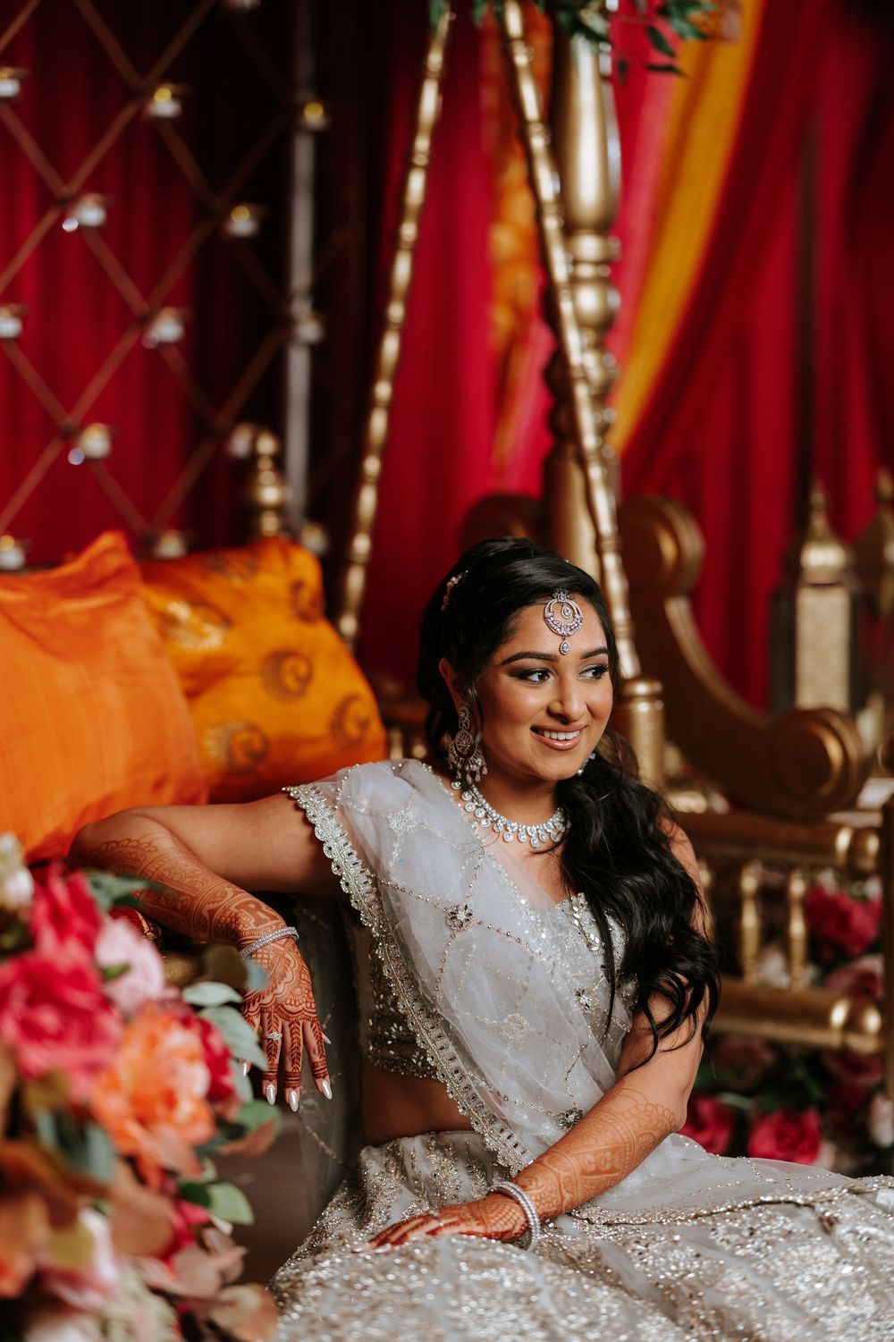 Bride in a sparkling white lehenga poses elegantly against a backdrop of rich red and gold decor.